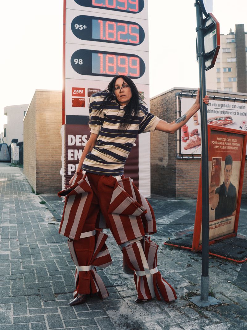 Model Boyd Gates posing in a striped top and sculptural striped trousers next to a gas station price display.