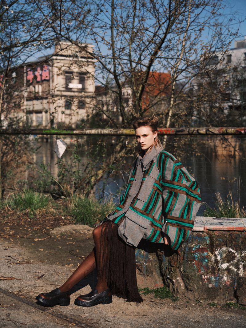 Model seated outdoors wearing a plaid jacket and dark shoes with bare branches and a building in the background