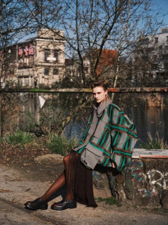 Model seated outdoors wearing a plaid jacket and dark shoes with bare branches and a building in the background