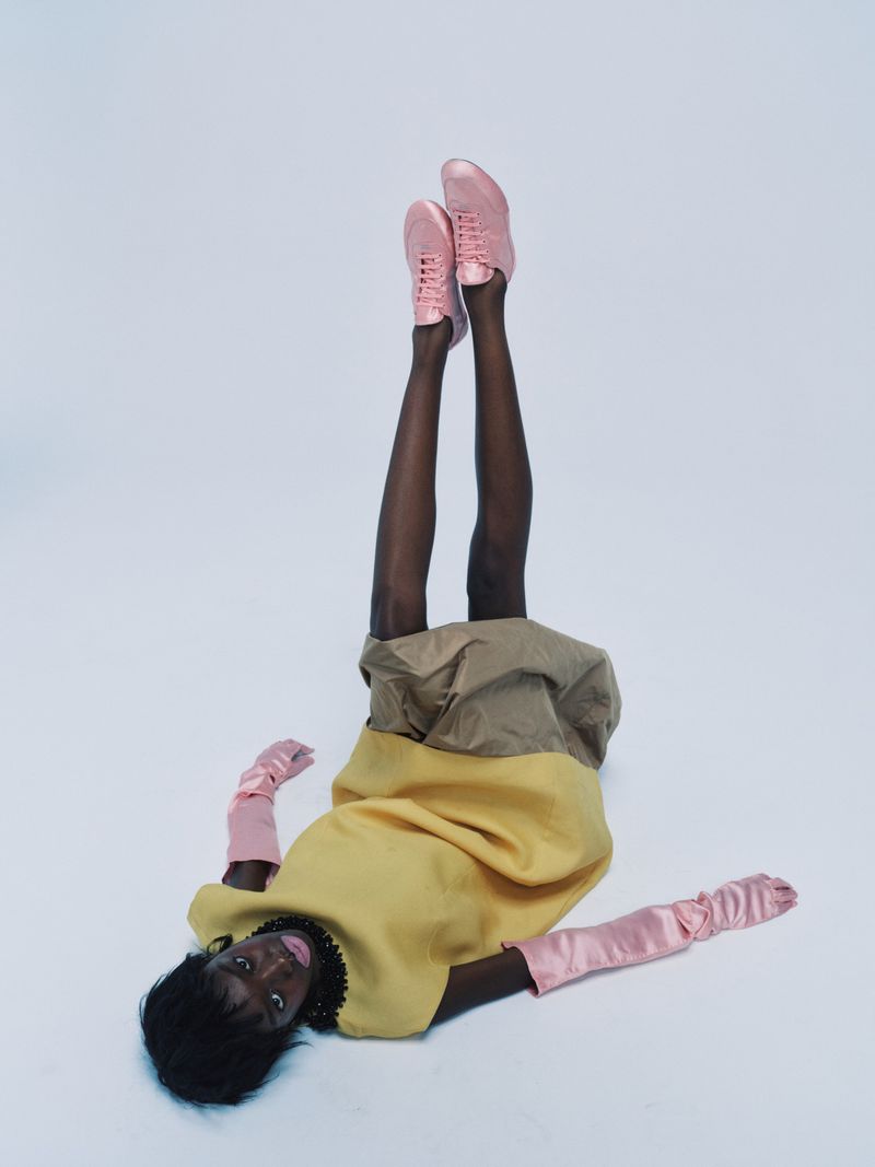 Model Boyd Gates lies on a studio floor with legs raised in the air wearing a yellow garment, pink gloves, and pink sneakers.