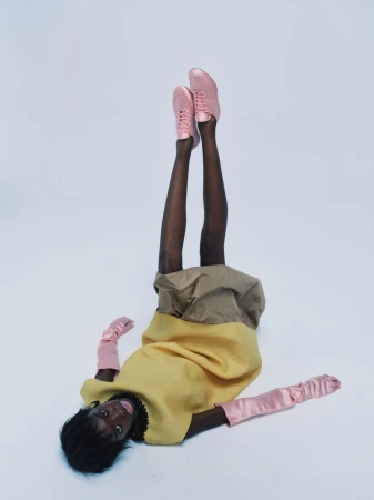 Model Boyd Gates lies on a studio floor with legs raised in the air wearing a yellow garment, pink gloves, and pink sneakers.