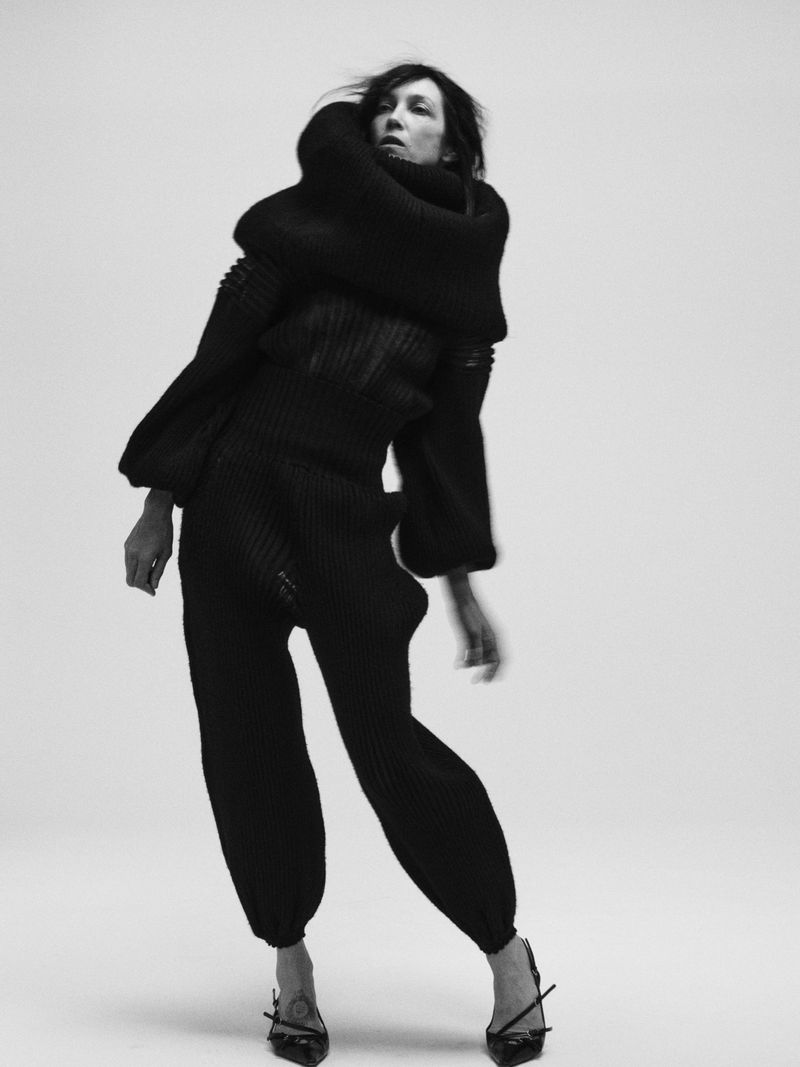 Model Boyd Gates posing in a sculptural black knit outfit with oversized cowl neck and pointed slingback heels against a white backdrop.