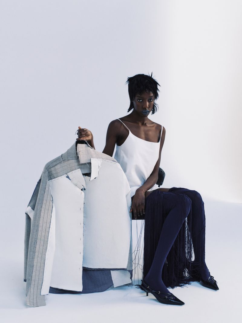 Model Boyd Gates seated in a white slip dress and dark tights holding a deconstructed structured jacket against a white studio background.