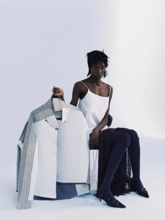 Model Boyd Gates seated in a white slip dress and dark tights holding a deconstructed structured jacket against a white studio background.