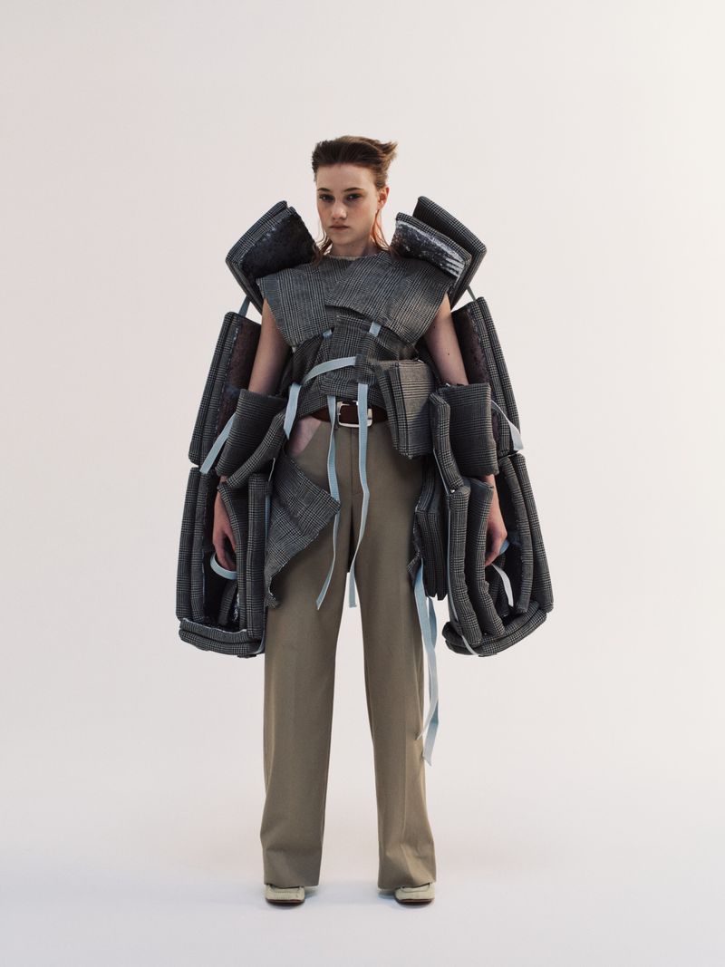 Model Boyd Gates posing in a sculptural grey panelled top and taupe wide leg trousers against a white seamless background.
