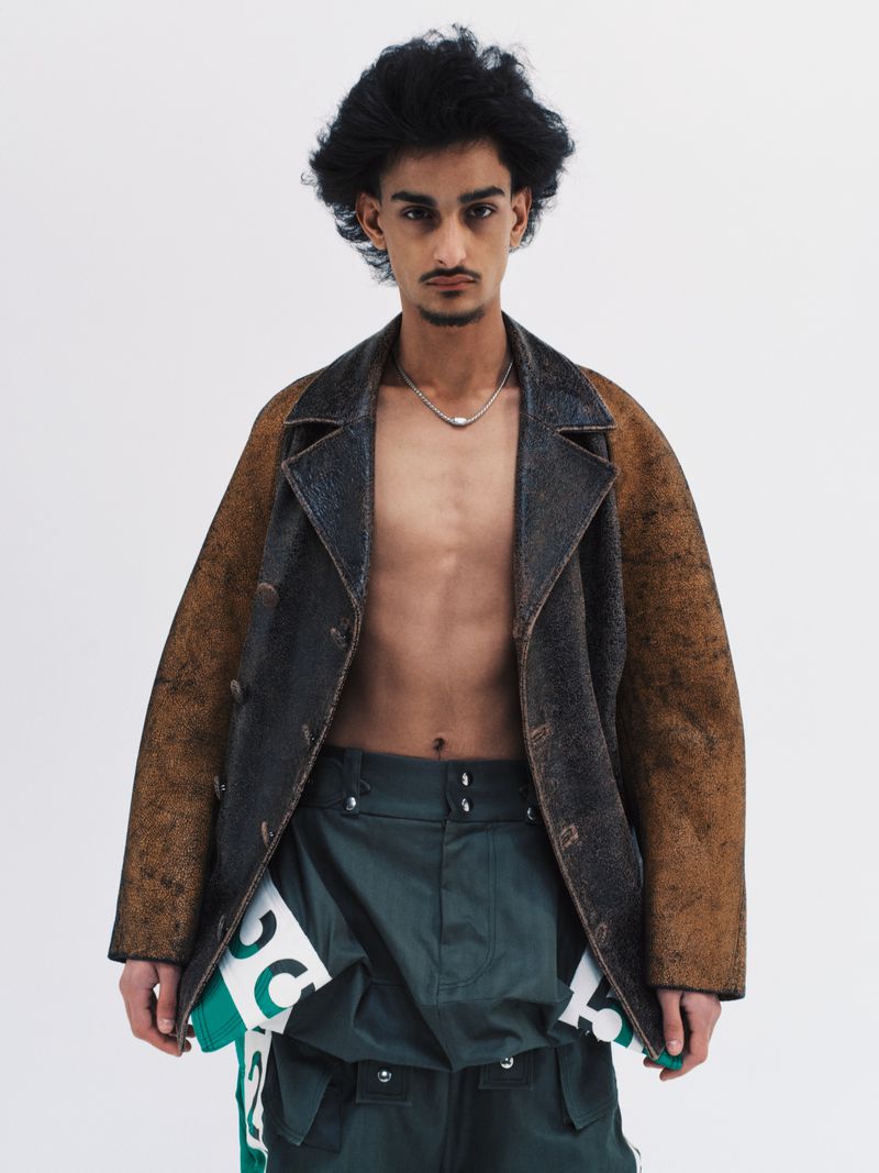 Boyd Gates wearing an open distressed leather jacket and dark green utility trousers against a plain white studio background.
