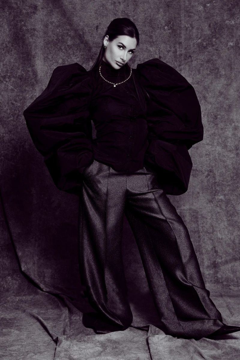 Model in an oversized black puff-sleeve top and dark wide-leg trousers posing against a mottled studio backdrop.