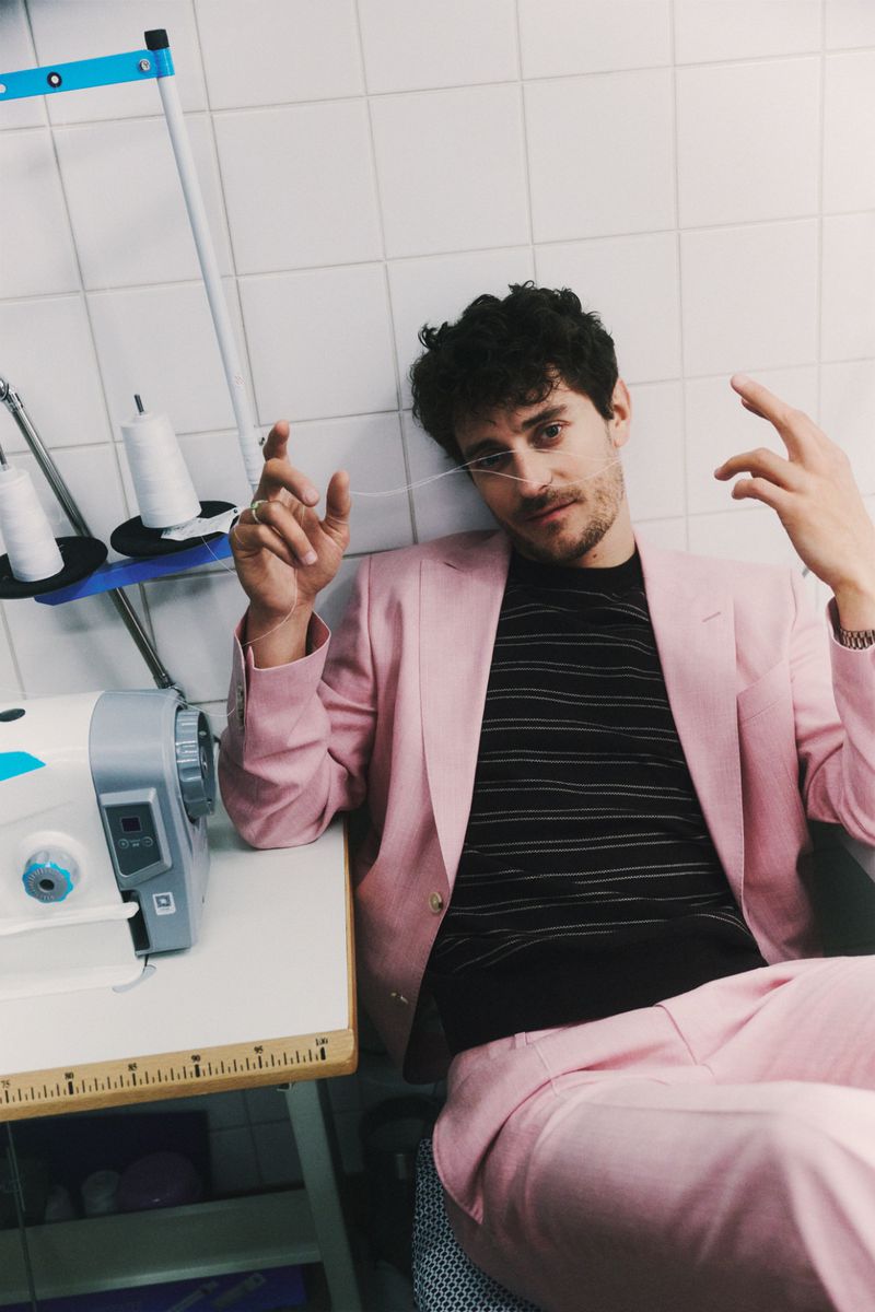 Jakub Józef Orliński wearing a tailored pink suit and black striped shirt, seated next to a sewing machine against a tiled wall.
