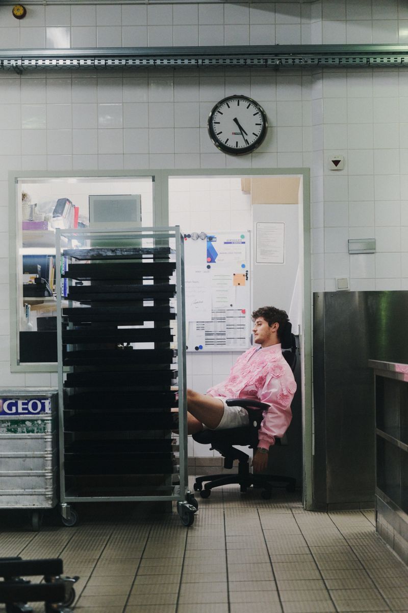 Man in a pink jacket seated in an office chair inside a commercial kitchen with tiled walls and a wall clock.