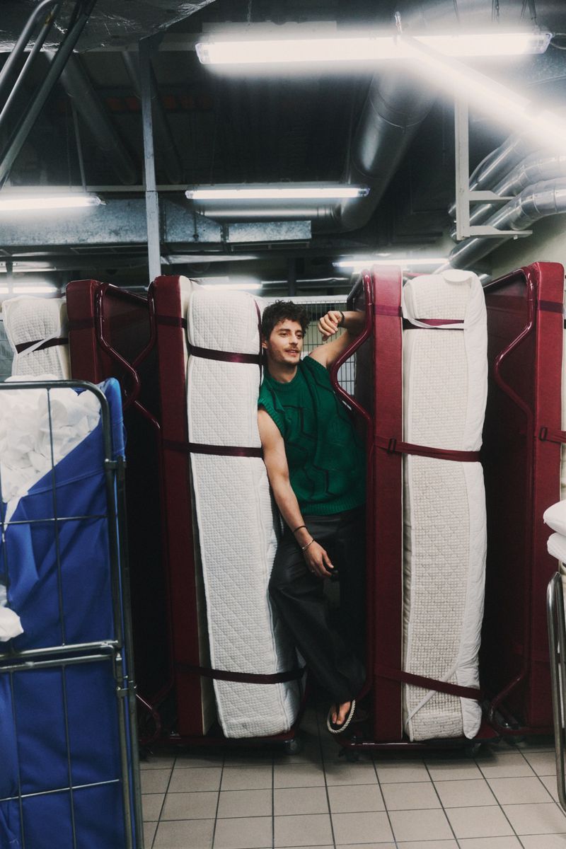 Jakub Józef Orliński posing between upright rollaway beds and mattresses in a green knit vest for GQ Poland.