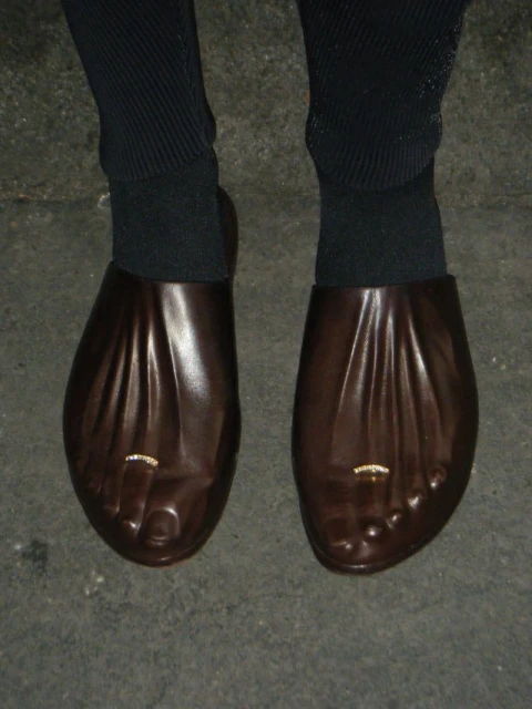 Unusual brown leather shoes shaped like bare feet with toe rings are worn with black socks on a grey concrete surface.