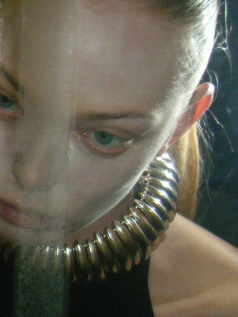 Close-up studio shot of a model wearing a large, sculptural gold choker necklace. The image has an artistic, slightly blurred aesthetic.