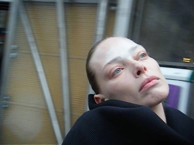 Close-up of a fair-skinned woman with light eyes looking upward, wearing black clothing, with a blurred background and pale makeup.
