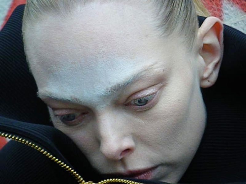 Close-up on a person with striking white face makeup and a black jacket featuring a gold zipper. The person's gaze is downward.