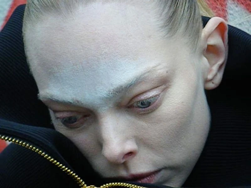 Close-up on a person with striking white face makeup and a black jacket featuring a gold zipper. The person's gaze is downward.