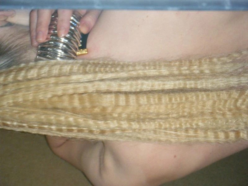 Close-up of crimped, blonde hair extensions in hand with metal hair crimper tool, captured indoors under ambient lighting.