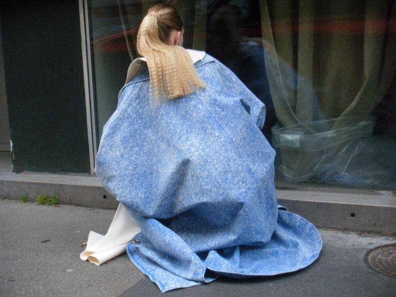 Back view of a person kneeling in front of a store window, wearing a long, light blue denim coat, with long blonde hair in a ponytail.