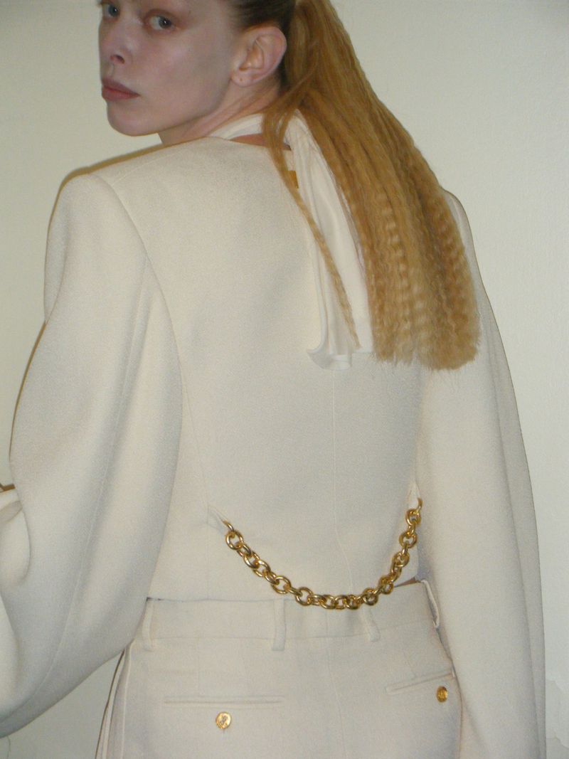 Back view of a fair-skinned adult woman wearing a white jacket and pants, with blonde crimped hair, and a gold chain on the pants.