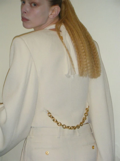 Back view of a fair-skinned adult woman wearing a white jacket and pants, with blonde crimped hair, and a gold chain on the pants.