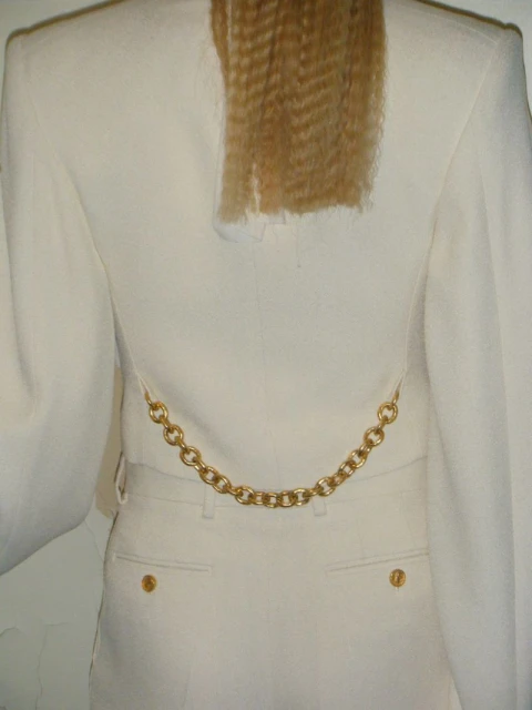 Back view of a person wearing a white suit jacket and pants, featuring a gold chain detail at the waist and crimped blonde hair.
