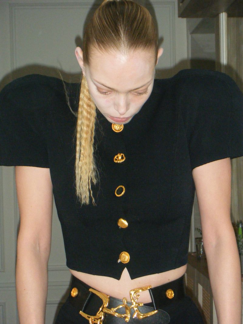 Model looking downward in a structured black crop top with exaggerated shoulders, gold buttons, and a decorative gold buckle belt.