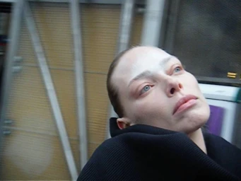 Close-up of a woman with slicked hair and pale makeup wearing a black ribbed sweater, tilting her head back against a blurred background.