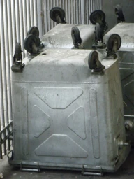 Large galvanized steel dumpsters turned upside down with caster wheels facing upright against vertical metal railings.