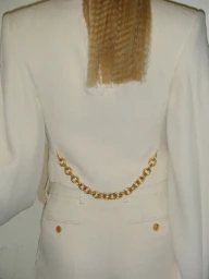 Back view of a model in a tailored cream suit jacket featuring a draped gold chain across the waistline and crimped blonde hair.