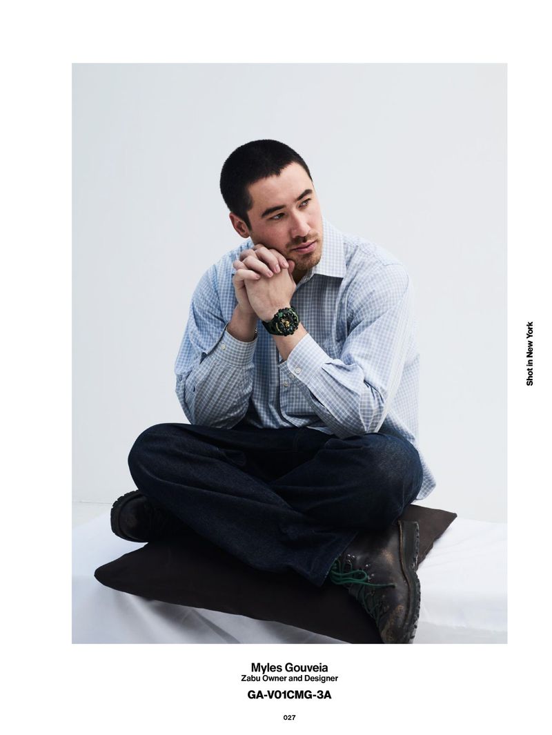 Man with short dark hair wearing a light blue patterned shirt and dark trousers sitting cross-legged against a white backdrop.