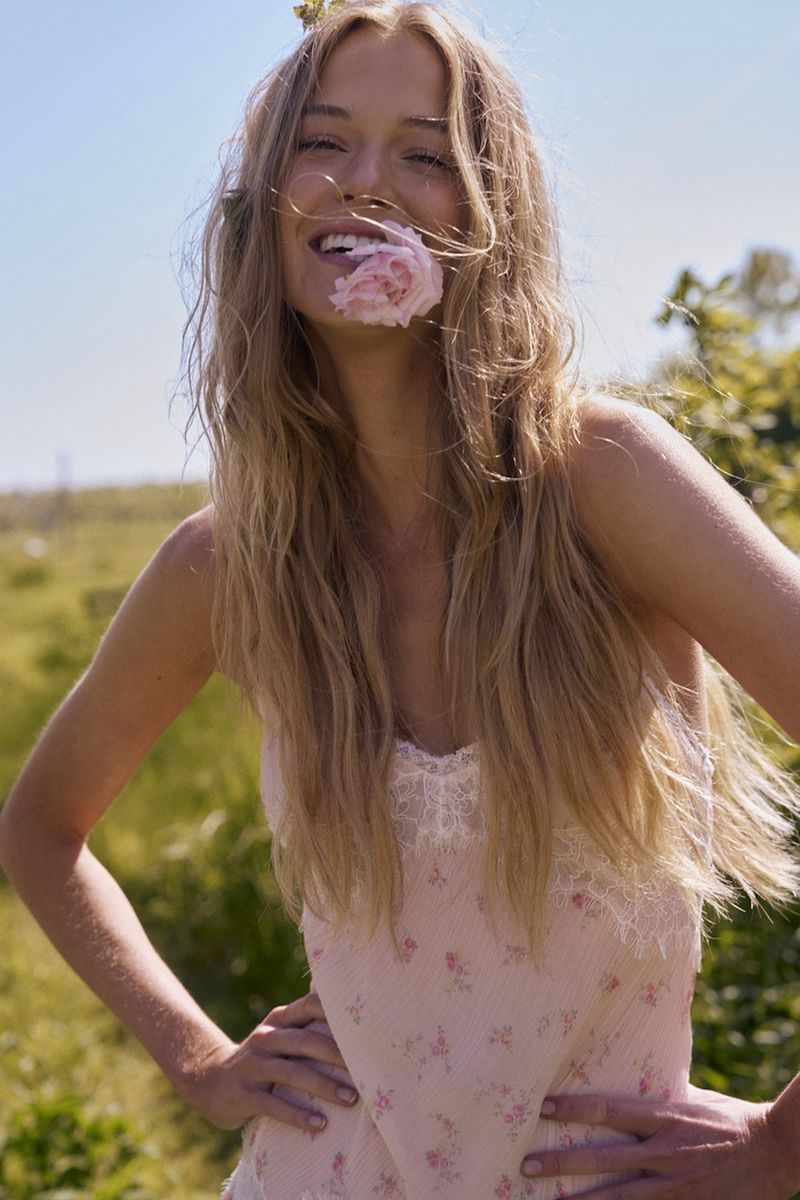 A smiling blonde woman holding a pink flower in her mouth, wearing a lace-trimmed floral camisole in a sunlit meadow.