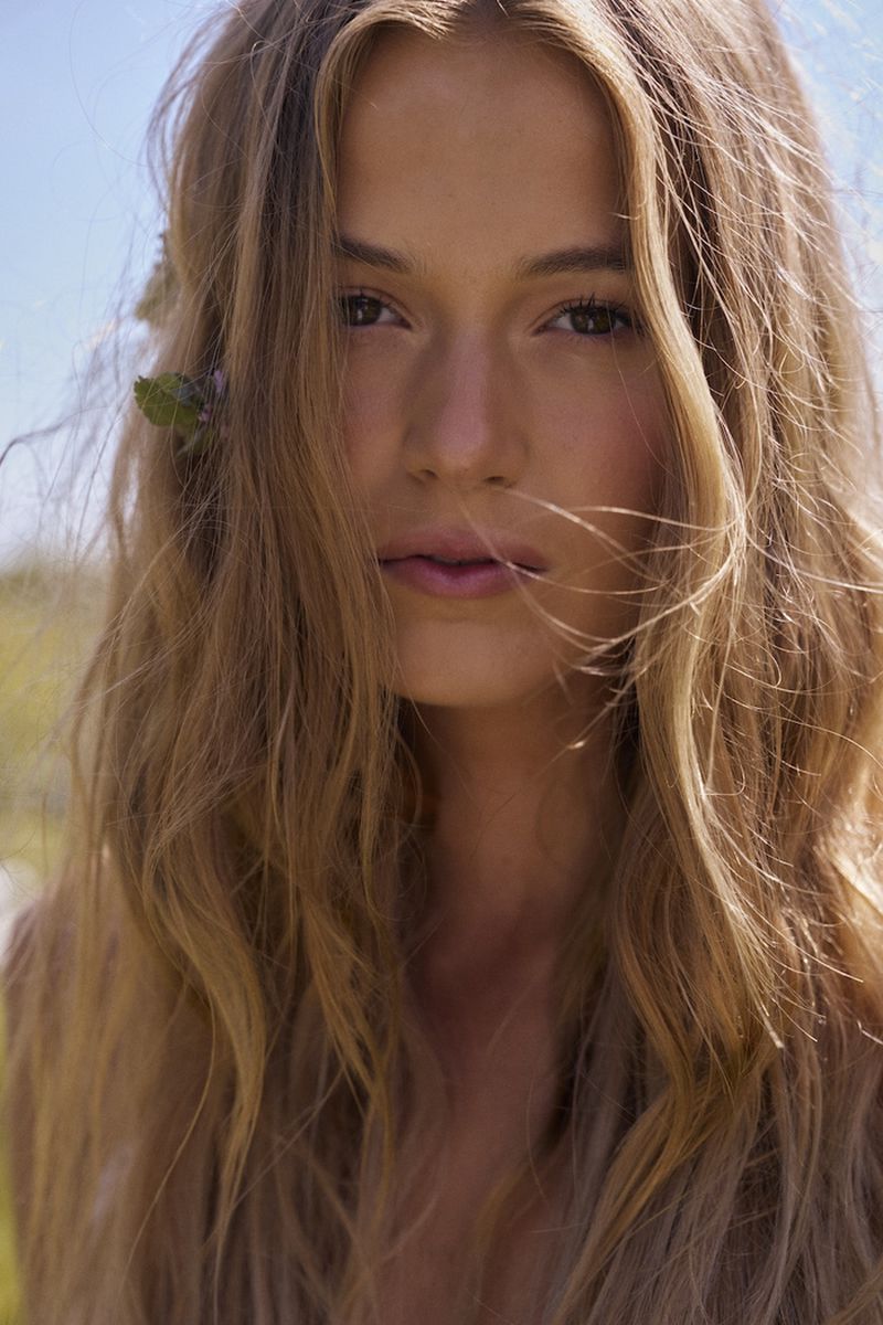 Close-up portrait of a woman with long wavy blonde hair and a small green plant tucked behind her ear against a bright sky.