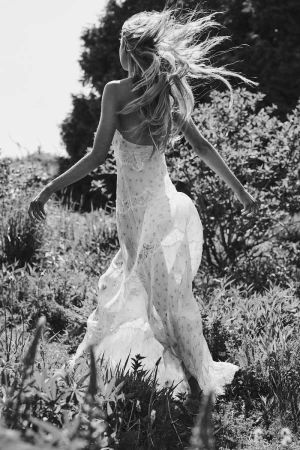 Monochrome view of a woman walking through a sunny meadow in a flowing lace dress with hair blowing in the wind.