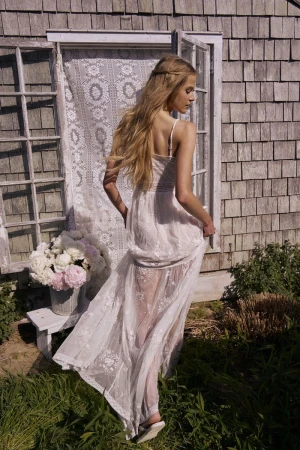 Woman in a sheer white embroidered lace maxi dress walking toward a rustic shingle cottage doorway.