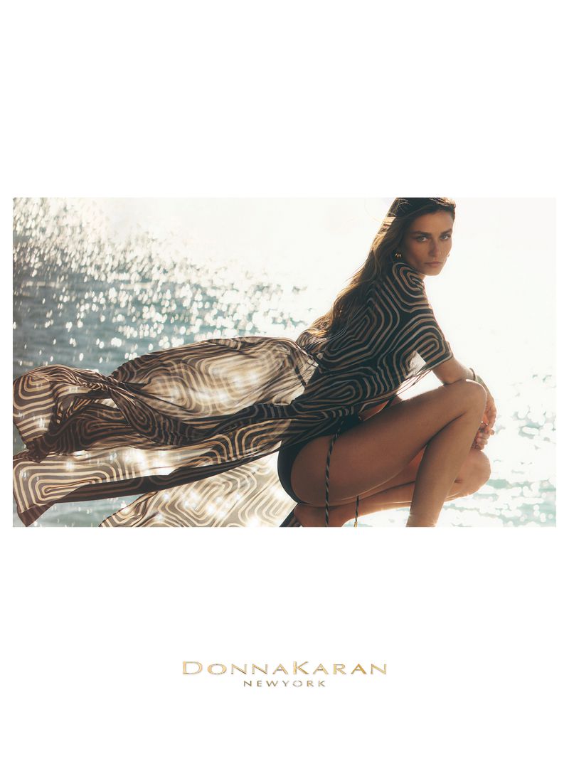 Model Andreea Diaconu wearing a flowing patterned sheer cover-up against sparkling sunlit water for Donna Karan.