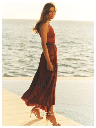 Model Andreea Diaconu posing in a pleated terracotta maxi dress and gold heeled sandals by the ocean for Donna Karan.