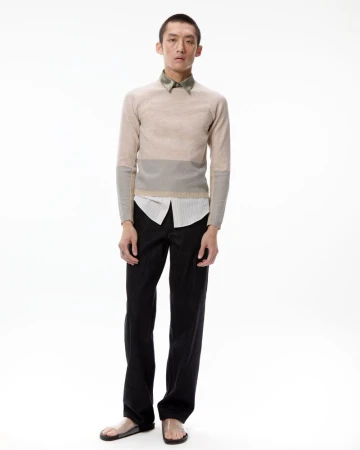 Model wearing a beige colorblock knit sweater, black trousers, and clear slide sandals against a white background.