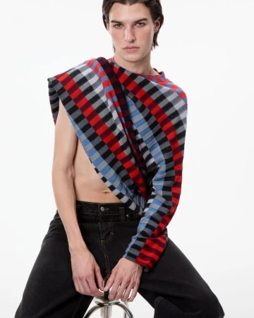 Model seated on a stool wearing an asymmetric checkered knit sweater and black denim against a white studio background.
