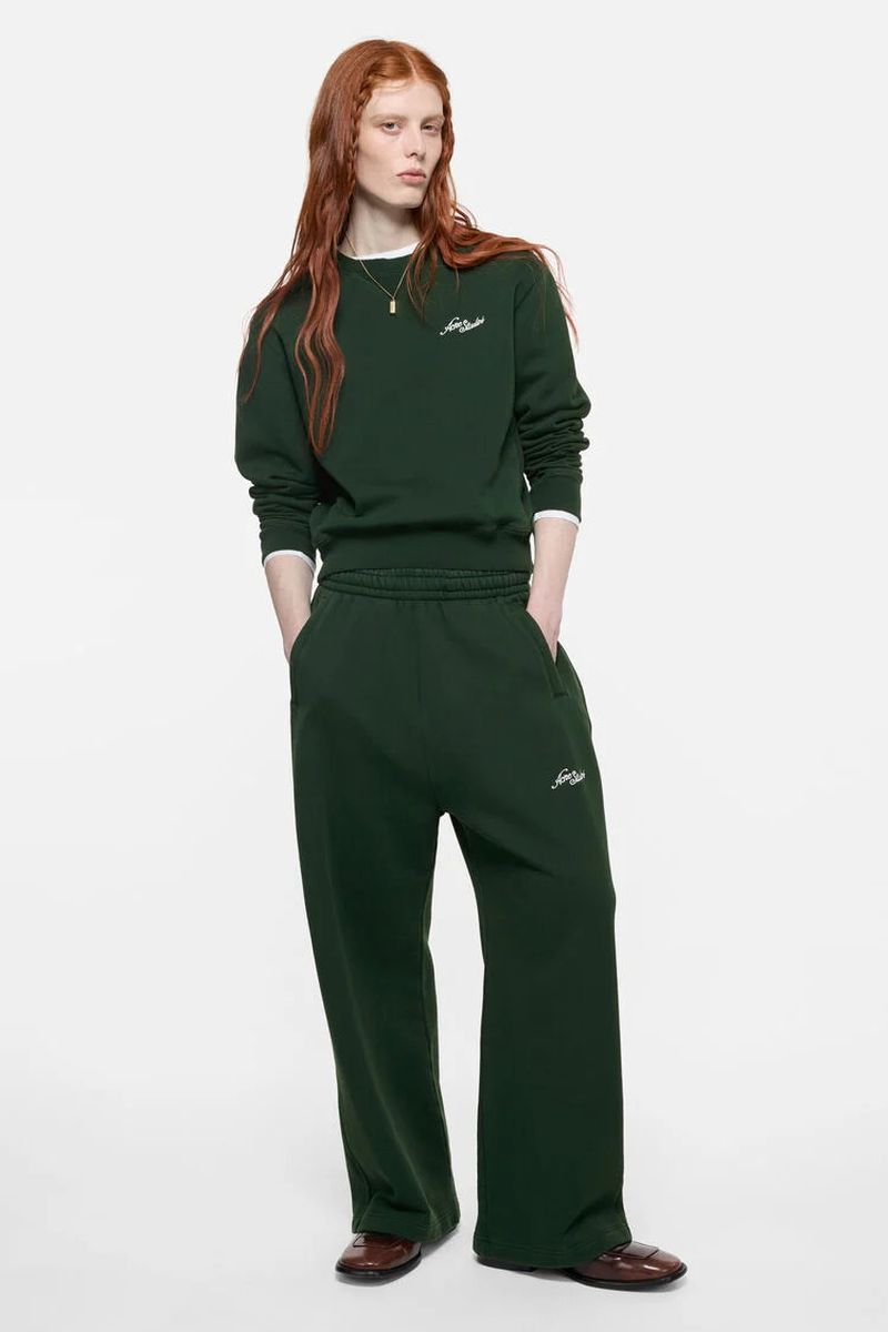 Model Laura Rudd wearing a dark forest green tracksuit with hands in pockets against a white seamless background.