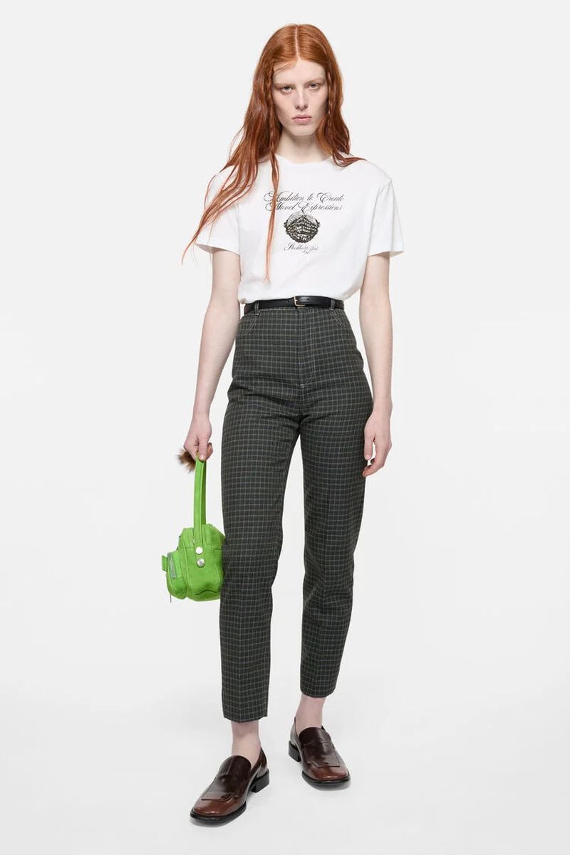 Model Laura Rudd wearing a white graphic t-shirt and grey plaid trousers holding a bright green bag against a white background.