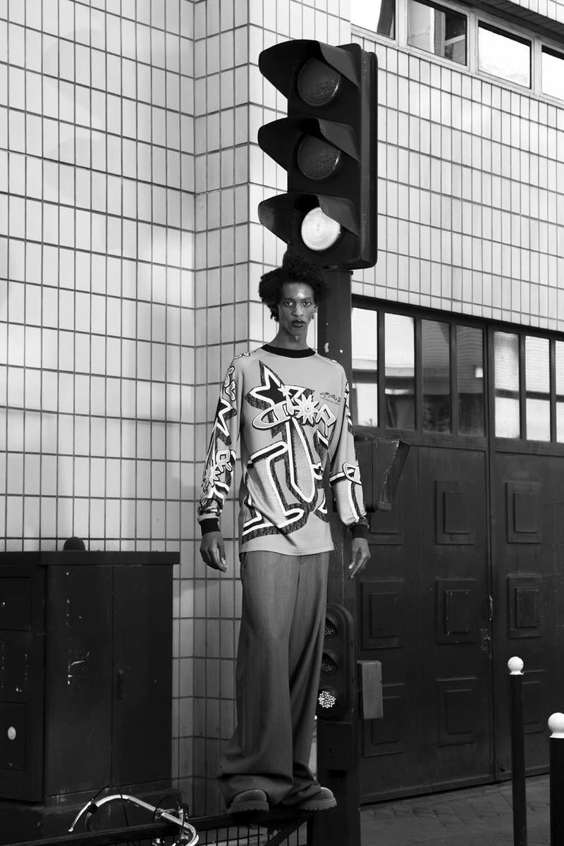 Black and white portrait of a person standing on a metal bar next to a traffic light and a tiled building in an urban setting.