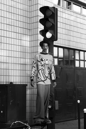 Black and white portrait of a person standing on a metal bar next to a traffic light and a tiled building in an urban setting.