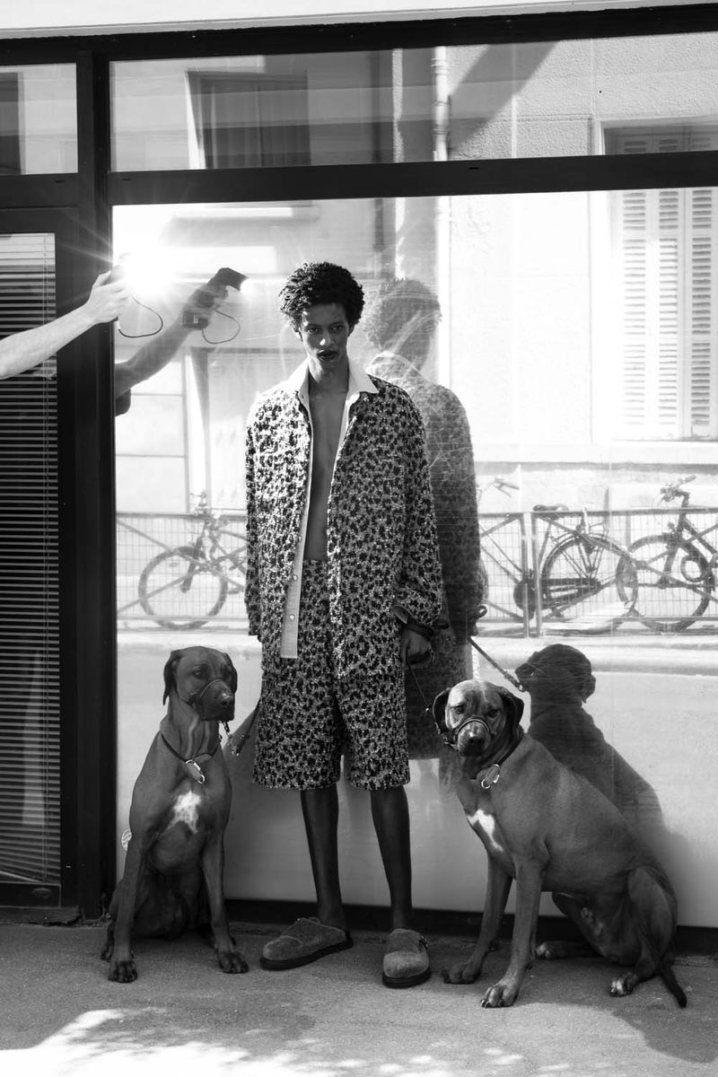 Fashion shot of a male model in a patterned outfit, posing with two dogs outdoors in front of a building on a sunny day.