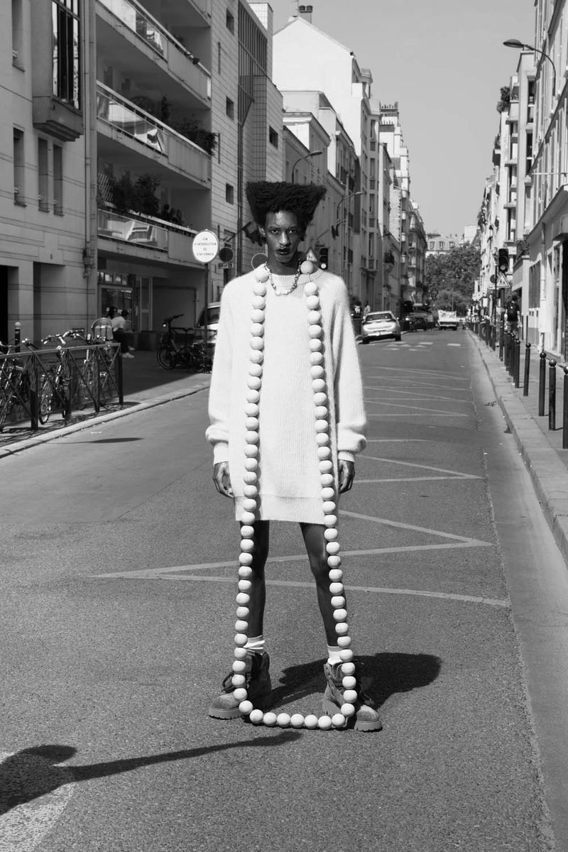 Craig Shimirimana wearing a knit sweater dress and floor-length spherical necklace on a city street in a black and white fashion portrait.