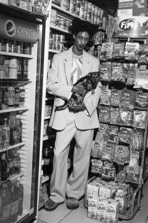 Black and white portrait of a Black male in a light suit holding a dog in a convenience store with shelves of candy and drinks.