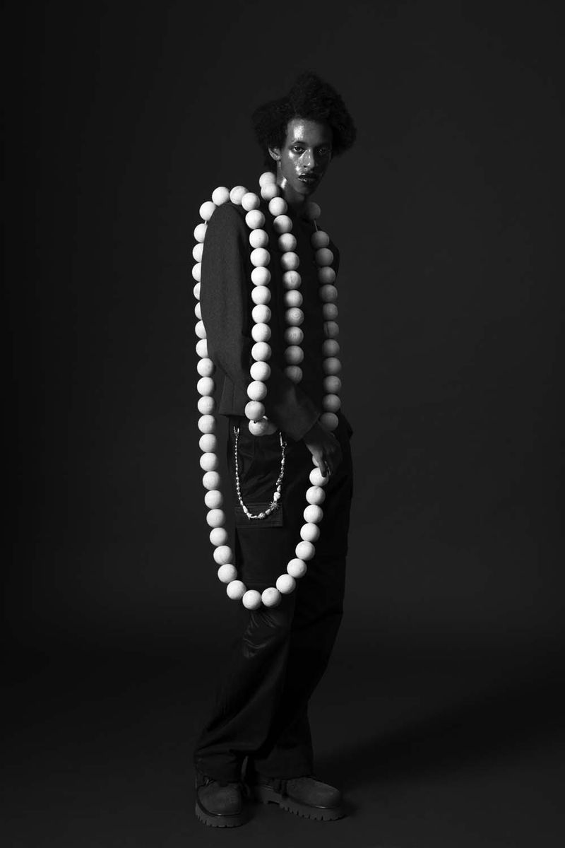 Black and white portrait of a man with an afro, wearing a black shirt and pants, and draped with large white spherical beads.