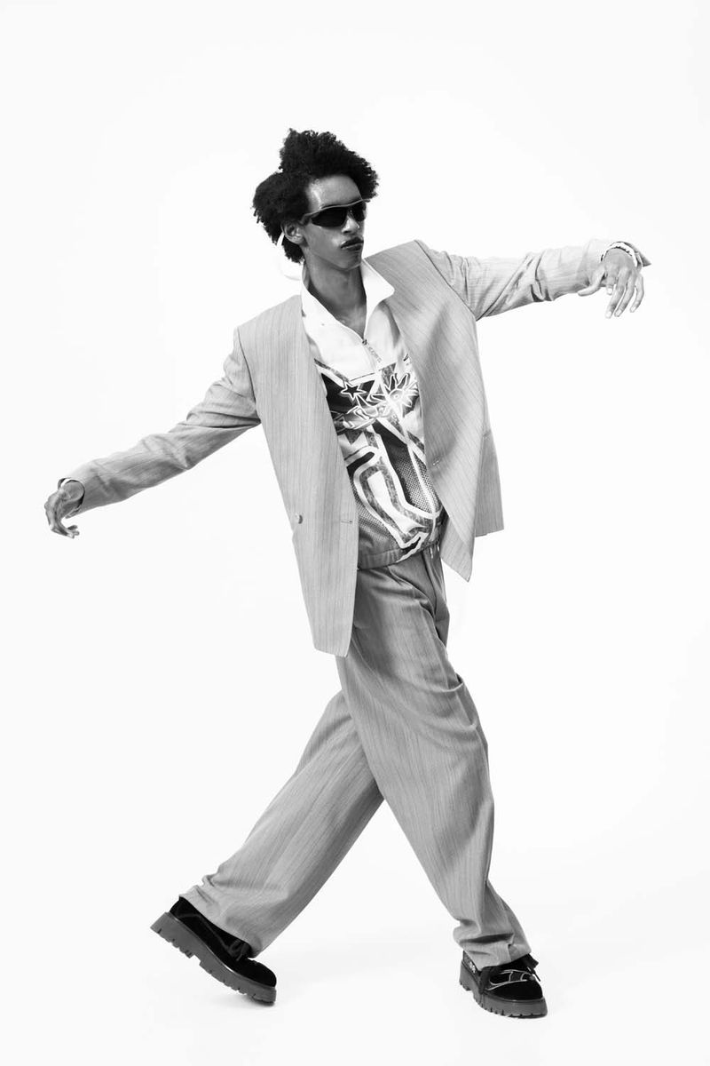 Black and white studio shot of a stylish African-American man in a suit, wide-legged pants, sunglasses, and loafers in a dynamic pose.