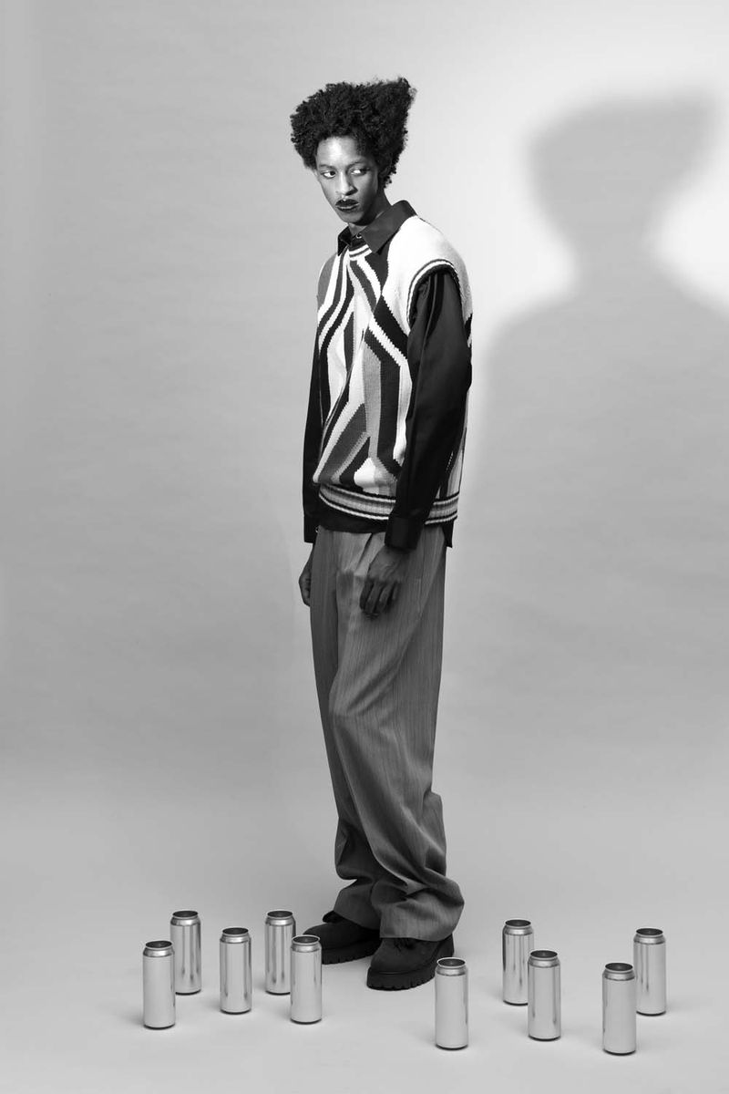 Black and white portrait of a slender adult man in a patterned sweater vest surrounded by aluminum cans in a grey photo studio.