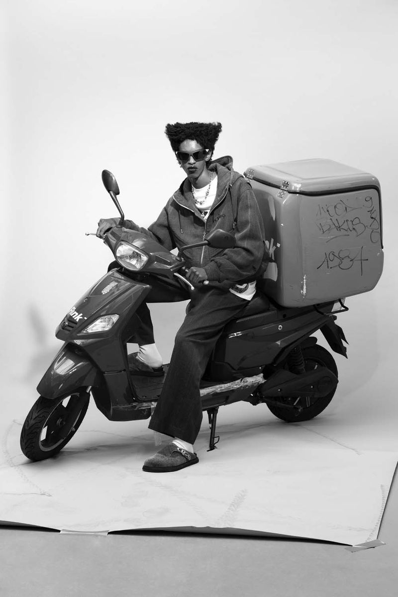 Person on a delivery scooter in a photo studio, wearing sunglasses and a hooded jacket, shot in black and white.