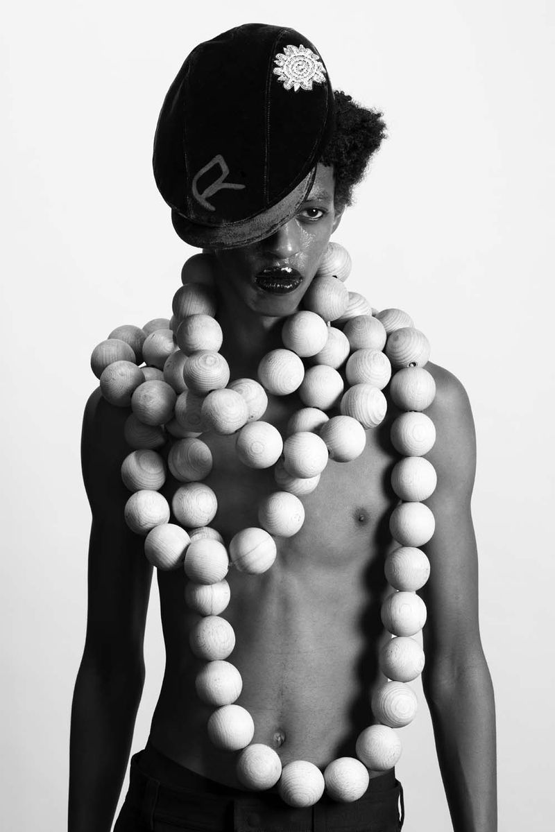 Monochrome portrait of a person with dark lipstick wearing a black cap, shirtless with beaded necklace, against a gradient background.