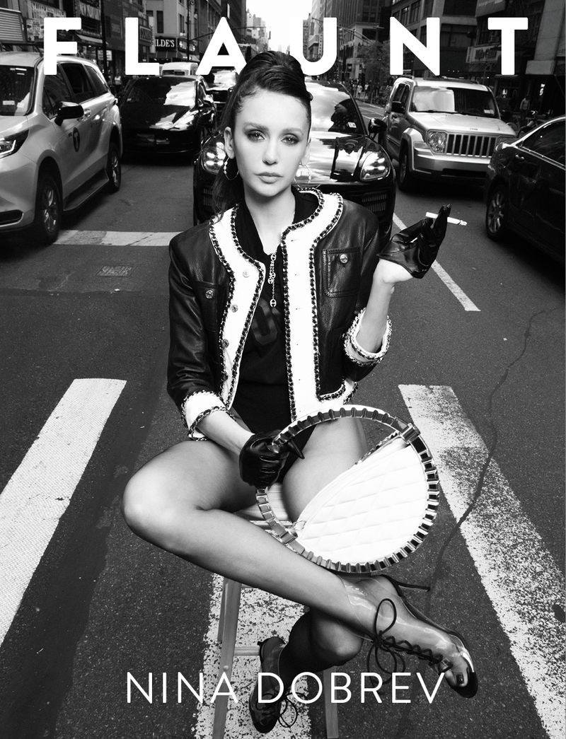 Black and white photo of Nina Dobrev seated on a street crosswalk in a trimmed jacket holding a bag for Flaunt Magazine.